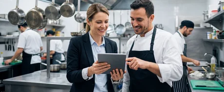 Austausch zwischen einer Frau und einem Mann in einer professionellen Großküche, die Frau hält ein Tablet in der Hand